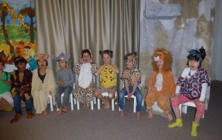 Children in different costumes sitting on chairs and giving a performance.
