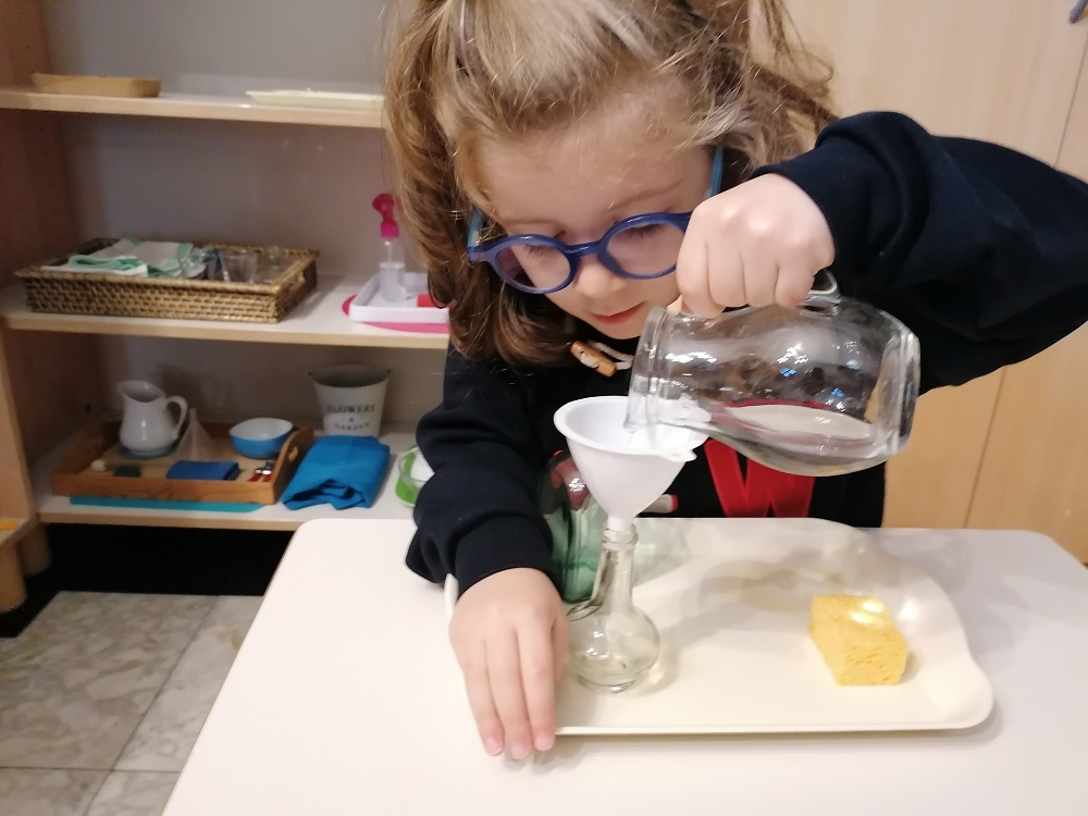 Girl in primary carefully pouring water from one jug into another using a beaker at Montessori House Brussels.